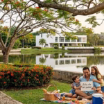 Una Fotografía Tomada En El Parque Tomás Garrido Canabal De Villahermosa, Tabasco. En El Primer Plano, A La Derecha, Un Hombre Sonriente Está Sentado Sobre Una Colorida Manta De Picnic En El Césped Junto A Sus Dos Hijos Pequeños; El Hombre Sostiene Un Letrero Blanco Que Dice &Quot;¡Feliz Día Del Padre!&Quot;. A Su Lado Hay Una Canasta De Picnic De Mimbre, Una Hielera Azul Y Un Perro Pequeño Sentado. A La Izquierda, Un Camino Empedrado Y Curvo Bordea Un Arbusto Con Flores Rojas Brillantes. El Fondo Muestra La Laguna De Las Ilusiones Reflejando Los Árboles Circundantes, Una Estructura Blanca Moderna Y El Emblemático Mirador Del Parque. Grandes Árboles Con Flores Rojas Enmarcan La Parte Superior De La Imagen.