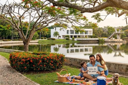 Una Fotografía Tomada En El Parque Tomás Garrido Canabal De Villahermosa, Tabasco. En El Primer Plano, A La Derecha, Un Hombre Sonriente Está Sentado Sobre Una Colorida Manta De Picnic En El Césped Junto A Sus Dos Hijos Pequeños; El Hombre Sostiene Un Letrero Blanco Que Dice &Quot;¡Feliz Día Del Padre!&Quot;. A Su Lado Hay Una Canasta De Picnic De Mimbre, Una Hielera Azul Y Un Perro Pequeño Sentado. A La Izquierda, Un Camino Empedrado Y Curvo Bordea Un Arbusto Con Flores Rojas Brillantes. El Fondo Muestra La Laguna De Las Ilusiones Reflejando Los Árboles Circundantes, Una Estructura Blanca Moderna Y El Emblemático Mirador Del Parque. Grandes Árboles Con Flores Rojas Enmarcan La Parte Superior De La Imagen.