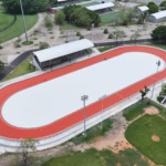 Vista Aérea Panorámica En Plano Picado Del Nuevo Patinódromo De Tabasco En El Deportivo Olimpia Xxi. La Instalación Deportiva Muestra Una Pista Ovalada De Patinaje Con Una Superficie Central Blanca Y Un Circuito Exterior De Color Rojo Brillante Perfectamente Delimitado. Junto A La Pista Se Observa Una Zona De Gradas Techada Con Una Estructura Arqueada Gris, Postes De Iluminación Profesional Y Áreas Verdes Circundantes Con Árboles Y Campos Deportivos Adyacentes.