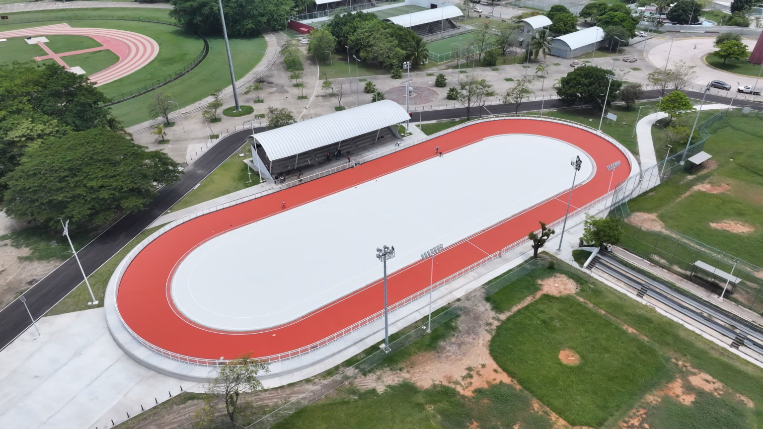 Vista Aérea Panorámica En Plano Picado Del Nuevo Patinódromo De Tabasco En El Deportivo Olimpia Xxi. La Instalación Deportiva Muestra Una Pista Ovalada De Patinaje Con Una Superficie Central Blanca Y Un Circuito Exterior De Color Rojo Brillante Perfectamente Delimitado. Junto A La Pista Se Observa Una Zona De Gradas Techada Con Una Estructura Arqueada Gris, Postes De Iluminación Profesional Y Áreas Verdes Circundantes Con Árboles Y Campos Deportivos Adyacentes.