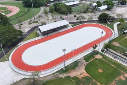 Vista Aérea Panorámica En Plano Picado Del Nuevo Patinódromo De Tabasco En El Deportivo Olimpia Xxi. La Instalación Deportiva Muestra Una Pista Ovalada De Patinaje Con Una Superficie Central Blanca Y Un Circuito Exterior De Color Rojo Brillante Perfectamente Delimitado. Junto A La Pista Se Observa Una Zona De Gradas Techada Con Una Estructura Arqueada Gris, Postes De Iluminación Profesional Y Áreas Verdes Circundantes Con Árboles Y Campos Deportivos Adyacentes.