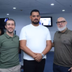 Fotografía De Plano Medio Que Muestra A Tres Hombres Sonriendo En El Centro De Un Salón Durante Un Evento Médico. A La Izquierda, Un Joven Viste Una Playera Tipo Polo Color Verde Oliva Y Pantalones Claros; En El Centro, Un Hombre Robusto Con Barba De Candado Viste Una Playera Blanca Y Mantiene Las Manos Cruzadas Al Frente; A La Derecha, Un Hombre Con Barba Clara, Lentes De Marco Negro Y Playera Tipo Polo Color Café Militar Complementa El Grupo.