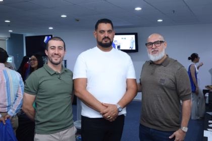 Fotografía De Plano Medio Que Muestra A Tres Hombres Sonriendo En El Centro De Un Salón Durante Un Evento Médico. A La Izquierda, Un Joven Viste Una Playera Tipo Polo Color Verde Oliva Y Pantalones Claros; En El Centro, Un Hombre Robusto Con Barba De Candado Viste Una Playera Blanca Y Mantiene Las Manos Cruzadas Al Frente; A La Derecha, Un Hombre Con Barba Clara, Lentes De Marco Negro Y Playera Tipo Polo Color Café Militar Complementa El Grupo.