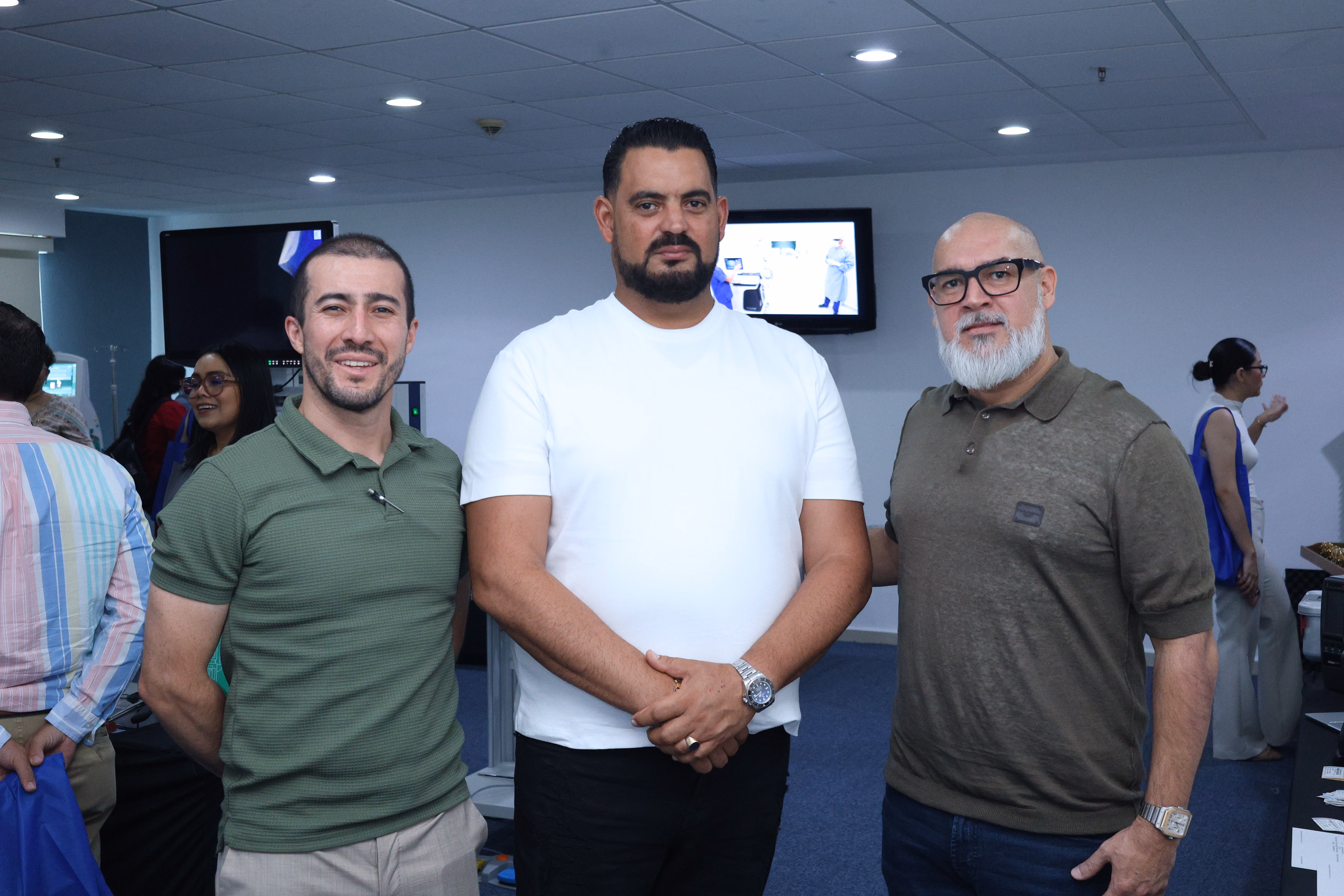 Fotografía De Plano Medio Que Muestra A Tres Hombres Sonriendo En El Centro De Un Salón Durante Un Evento Médico. A La Izquierda, Un Joven Viste Una Playera Tipo Polo Color Verde Oliva Y Pantalones Claros; En El Centro, Un Hombre Robusto Con Barba De Candado Viste Una Playera Blanca Y Mantiene Las Manos Cruzadas Al Frente; A La Derecha, Un Hombre Con Barba Clara, Lentes De Marco Negro Y Playera Tipo Polo Color Café Militar Complementa El Grupo.