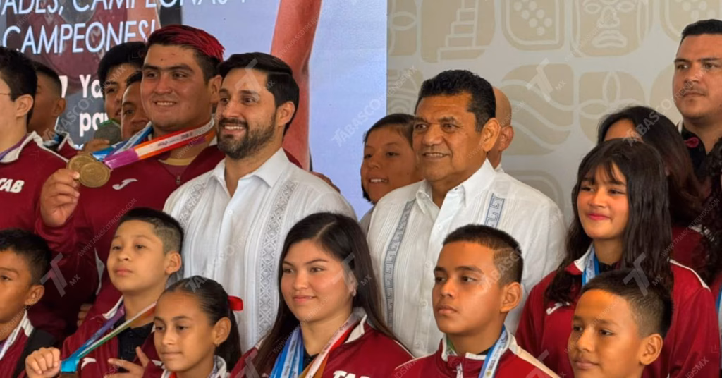 Javier May Presenta Logros En Deporte Y Transparencia En Tabasco 71 Fotografía En Plano Medio Y Encuadre Horizontal De Un Grupo De Atletas Infantiles Y Juveniles Posando Junto A Autoridades. Al Fondo A La Derecha, Destaca El Gobernador De Tabasco Portando Una Guayabera Blanca De Manga Larga Con Alforzas Y Bordados Geométricos Claros. A Su Lado Izquierdo Se Encuentra Otro Funcionario Con Una Prenda Similar. Los Jóvenes Deportistas Rodean A Las Autoridades Vistiendo Chamarras Deportivas De Color Guinda Con Franjas Blancas En Los Hombros, Correspondientes Al Uniforme Oficial Del Estado.