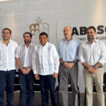 Fotografía De Plano Medio Que Muestra A Cinco Hombres De Pie, Alineados Horizontalmente Durante Un Evento Oficial En Tabasco. En El Centro Se Encuentra El Gobernador De Tabasco, Javier May Rodríguez, Vistiendo Una Guayabera Blanca Con Bordados Tradicionales En Color Azul Y Pantalón Oscuro. A Su Izquierda (Derecha De La Imagen), Lo Acompañan Dos Empresarios: Uno Con Camisa De Vestir Azul Claro Y Pantalón Gris, Y Otro Con Camisa A Cuadros Claros Y Pantalón Beige, Ambos Con Las Manos Entrelazadas Al Frente