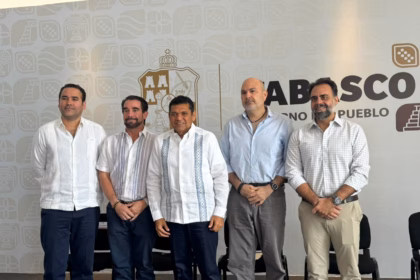 Fotografía De Plano Medio Que Muestra A Cinco Hombres De Pie, Alineados Horizontalmente Durante Un Evento Oficial En Tabasco. En El Centro Se Encuentra El Gobernador De Tabasco, Javier May Rodríguez, Vistiendo Una Guayabera Blanca Con Bordados Tradicionales En Color Azul Y Pantalón Oscuro. A Su Izquierda (Derecha De La Imagen), Lo Acompañan Dos Empresarios: Uno Con Camisa De Vestir Azul Claro Y Pantalón Gris, Y Otro Con Camisa A Cuadros Claros Y Pantalón Beige, Ambos Con Las Manos Entrelazadas Al Frente