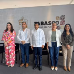 Fotografía En Plano Entero De Cinco Funcionarios Públicos De Pie Y Alineados De Izquierda A Derecha En Un Foro Oficial. Al Centro Se Encuentra El Gobernador De Tabasco, Vistiendo Una Guayabera Blanca Tradicional De Manga Larga Y Pantalón Oscuro. A Su Izquierda, Un Hombre Con Guayabera Blanca Con Bordados Azules Y Pantalón De Mezclilla; En El Extremo Izquierdo, Una Mujer Con Un Conjunto De Dos Piezas Con Estampado Floral En Tonos Rosa Y Naranja.