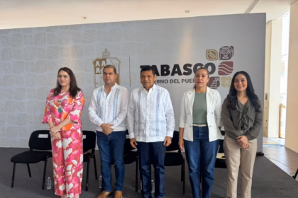 Fotografía En Plano Entero De Cinco Funcionarios Públicos De Pie Y Alineados De Izquierda A Derecha En Un Foro Oficial. Al Centro Se Encuentra El Gobernador De Tabasco, Vistiendo Una Guayabera Blanca Tradicional De Manga Larga Y Pantalón Oscuro. A Su Izquierda, Un Hombre Con Guayabera Blanca Con Bordados Azules Y Pantalón De Mezclilla; En El Extremo Izquierdo, Una Mujer Con Un Conjunto De Dos Piezas Con Estampado Floral En Tonos Rosa Y Naranja.