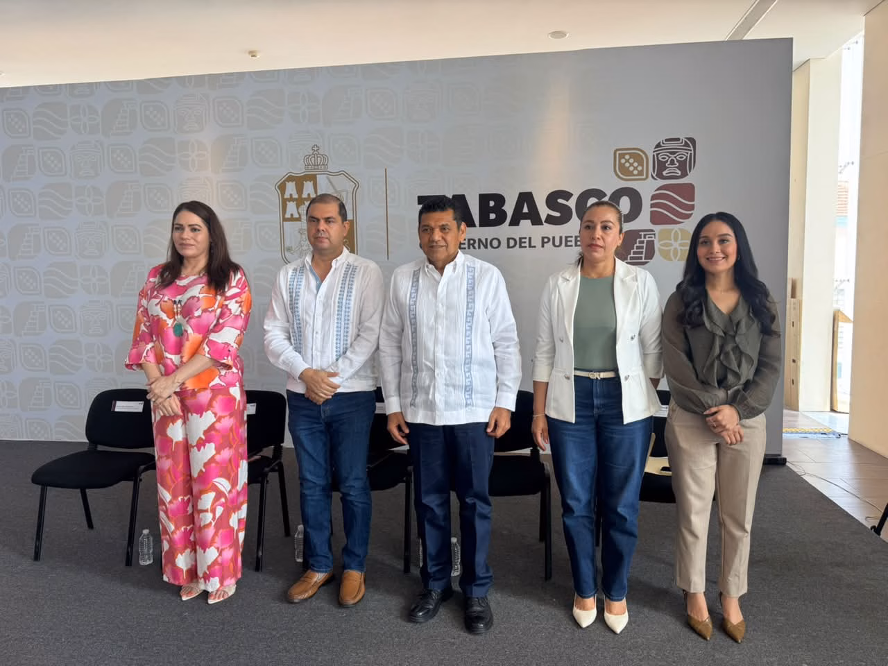 Javier May Presenta Logros En Deporte Y Transparencia En Tabasco 70 Fotografía En Plano Entero De Cinco Funcionarios Públicos De Pie Y Alineados De Izquierda A Derecha En Un Foro Oficial. Al Centro Se Encuentra El Gobernador De Tabasco, Vistiendo Una Guayabera Blanca Tradicional De Manga Larga Y Pantalón Oscuro. A Su Izquierda, Un Hombre Con Guayabera Blanca Con Bordados Azules Y Pantalón De Mezclilla; En El Extremo Izquierdo, Una Mujer Con Un Conjunto De Dos Piezas Con Estampado Floral En Tonos Rosa Y Naranja.