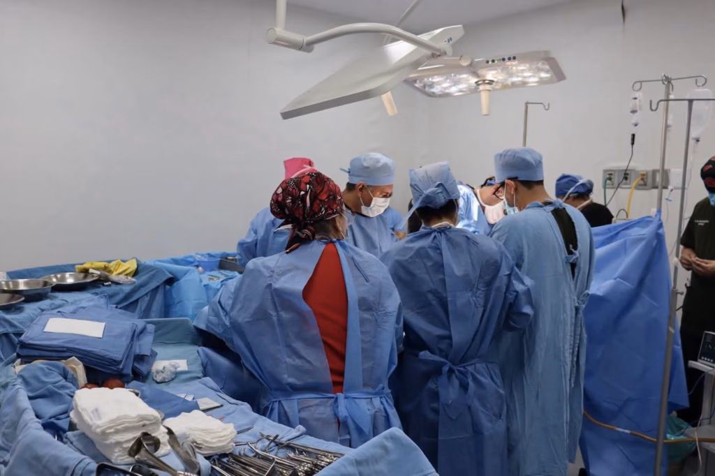Fotografía Horizontal En Plano Medio Que Muestra El Interior De Un Quirófano Iluminado Por Una Lámpara Quirúrgica De Techo. Un Equipo Multidisciplinario De Cirujanos Y Enfermeros Especialistas, Vestidos Con Batas Azules, Gorros Quirúrgicos, Cubrebocas Y Guantes, Trabaja De Manera Coordinada Alrededor De La Mesa De Operaciones