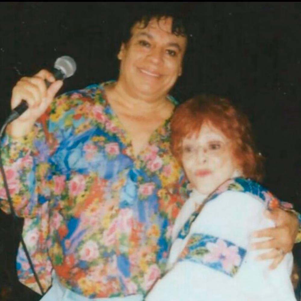 Se Observa Una Fotografía Vintage De Los Reconocidos Cantantes Mexicanos Juan Gabriel Y Dora María Posando Juntos De Medio Cuerpo Sobre Un Fondo Oscuro. A La Izquierda, Un Joven Juan Gabriel Sonríe De Frente A La Cámara Mientras Sostiene Un Micrófono Con La Mano Derecha Alzada; Viste Una Camisa Holgada De Manga Larga Con Un Llamativo Estampado Floral Multicolor. Con Su Brazo Izquierdo, Abraza Cariñosamente Por El Hombro A Dora María &Quot;La Chaparrita De Oro&Quot;,