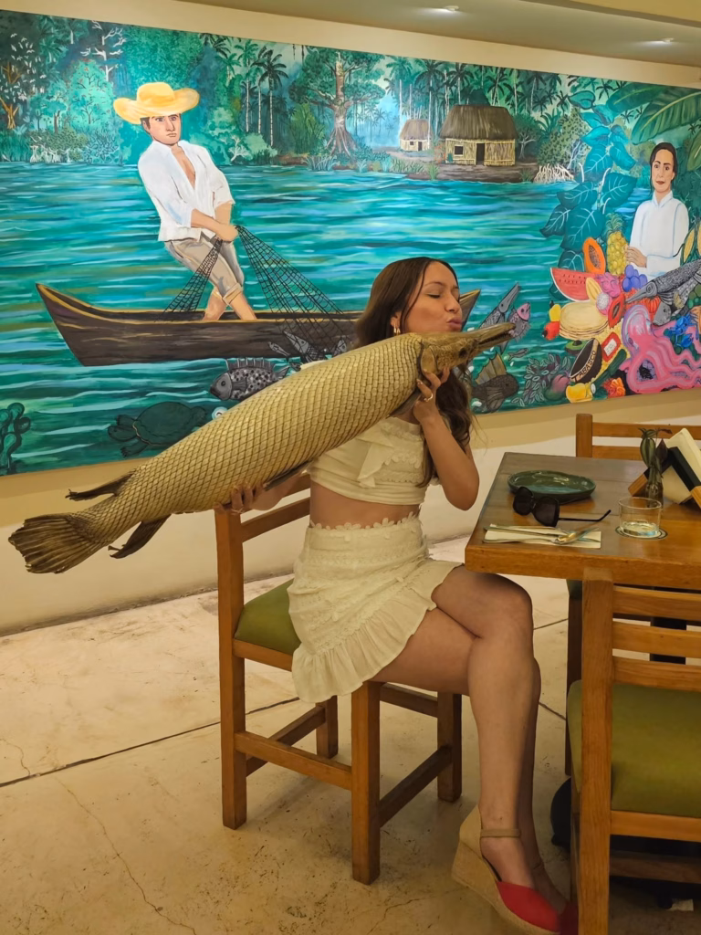 ¡Tabasco Es Un Edén! La Famosa Tiktoker &Quot;Miss Azalhia&Quot; Enamora A Sus Seguidores Con Su Viaje Por Villahermosa 71 Fotografía De La Creadora De Contenido Azalhia Edith Sentada En Un Restaurante, Vistiendo Un Conjunto De Dos Piezas Blanco Y Zapatillas De Plataforma. Sostiene Con Ambas Manos Un Pejelagarto Entero Disecado Y Simula Darle Un Beso. Al Fondo, Destaca Un Gran Mural Colorido Con Paisajes Tropicales De Tabasco, Pescadores Y Motivos Regionales.