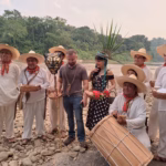 Una Fotografía Al Aire Libre Junto Al Río En El Pueblo Mágico De Tapijulapa, Tabasco, Donde La Actriz Maribel Guardia Posa Junto A Un Grupo De Música Y Danza Tradicional. Maribel Guardia Viste Un Vestido Negro Con Lunares Blancos Y Un Tocado Artesanal De Plumas Verdes Y Cuentas Azules Sobre Su Cabeza, Mientras Sostiene Una Pequeña Canasta Con Flores Rojas. A Su Alrededor, Varios Hombres Vestidos Con La Indumentaria Tradicional Tabasqueña De Manta Blanca, Pañuelos Rojos Al Cuello Y Sombreros De Chontalpa Sostienen Cestos De Mimbre, Velas Y Un Tambor Tradicional. Un Hombre Con Playera Gris Y Jeans Se Encuentra De Pie Junto A Ella En El Centro. El Grupo Está Posando Sobre Una Orilla Llena De Piedras De Río Redondas, Con Agua Mansa Y Una Densa Vegetación Selvática De Fondo Bajo Un Cielo Claro.