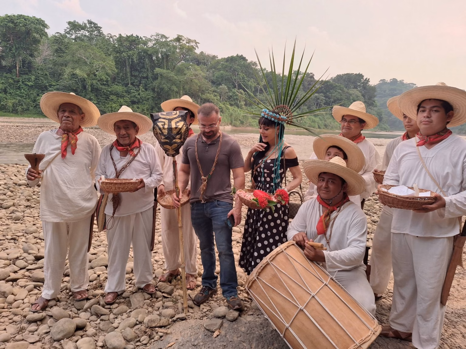 Maribel Guardia Enamora A Tacotalpa Y Baila Al Ritmo Del Zapateado En Tapijulapa 69 Una Fotografía Al Aire Libre Junto Al Río En El Pueblo Mágico De Tapijulapa, Tabasco, Donde La Actriz Maribel Guardia Posa Junto A Un Grupo De Música Y Danza Tradicional. Maribel Guardia Viste Un Vestido Negro Con Lunares Blancos Y Un Tocado Artesanal De Plumas Verdes Y Cuentas Azules Sobre Su Cabeza, Mientras Sostiene Una Pequeña Canasta Con Flores Rojas. A Su Alrededor, Varios Hombres Vestidos Con La Indumentaria Tradicional Tabasqueña De Manta Blanca, Pañuelos Rojos Al Cuello Y Sombreros De Chontalpa Sostienen Cestos De Mimbre, Velas Y Un Tambor Tradicional. Un Hombre Con Playera Gris Y Jeans Se Encuentra De Pie Junto A Ella En El Centro. El Grupo Está Posando Sobre Una Orilla Llena De Piedras De Río Redondas, Con Agua Mansa Y Una Densa Vegetación Selvática De Fondo Bajo Un Cielo Claro.