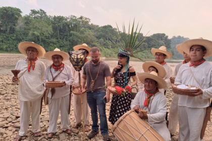 Una Fotografía Al Aire Libre Junto Al Río En El Pueblo Mágico De Tapijulapa, Tabasco, Donde La Actriz Maribel Guardia Posa Junto A Un Grupo De Música Y Danza Tradicional. Maribel Guardia Viste Un Vestido Negro Con Lunares Blancos Y Un Tocado Artesanal De Plumas Verdes Y Cuentas Azules Sobre Su Cabeza, Mientras Sostiene Una Pequeña Canasta Con Flores Rojas. A Su Alrededor, Varios Hombres Vestidos Con La Indumentaria Tradicional Tabasqueña De Manta Blanca, Pañuelos Rojos Al Cuello Y Sombreros De Chontalpa Sostienen Cestos De Mimbre, Velas Y Un Tambor Tradicional. Un Hombre Con Playera Gris Y Jeans Se Encuentra De Pie Junto A Ella En El Centro. El Grupo Está Posando Sobre Una Orilla Llena De Piedras De Río Redondas, Con Agua Mansa Y Una Densa Vegetación Selvática De Fondo Bajo Un Cielo Claro.