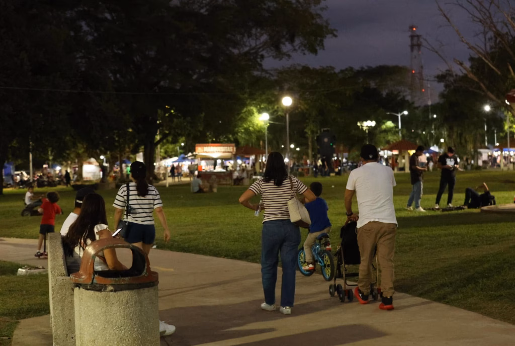 Guía Para El Día Del Padre 2026: Lugares Gratis En Villahermosa Para Un Día De Campo Inolvidable 3 Fotografía Nocturna En Un Plano Medio Que Muestra El Ambiente Familiar En El Parque La Choca En Villahermosa, Tabasco. Varias Personas Caminan Por Un Sendero Pavimentado Que Atraviesa Una Amplia Zona De Césped Iluminada Por Faroles Públicos. En Primer Plano, De Espaldas, Se Observa A Una Mujer Con Playera A Rayas Y Jeans Junto A Un Hombre Con Playera Blanca Y Pantalón Café Que Empuja Una Carriola; A Su Lado, Un Niño Pequeño Va En Una Bicicleta Azul. A La Izquierda, Una Mujer Está Sentada En Una Banca De Concreto Junto A Un Bote De Basura, Y Al Fondo Se Distinguen Árboles, Puestos De Comida Iluminados Y Otras Personas Disfrutando De La Tarde.