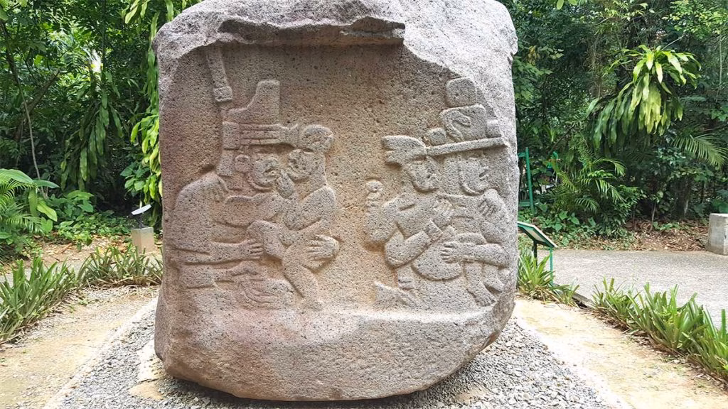 Un Gran Monumento Prehispánico De Piedra Con Relieves Tallados Que Muestran Figuras Humanas De Perfil Con Tocados Y Vestimentas De La Cultura Olmeca, Ubicado En Un Sendero Al Aire Libre Rodeado De Selva Tropical.