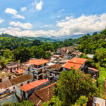 Vista Panorámica Desde Las Alturas Del Pueblo Mágico De Tapijulapa, Tabasco, Mostrando Un Pintoresco Conjunto De Casas Con Tejados De Teja De Arcilla Marrón Y Techos Rojizos, Enclavadas En Un Valle Rodeado Por Exuberantes Montañas Y Cerros Cubiertos De Densa Vegetación Selvática. Un Cielo Azul Brillante Salpicado De Nubes Blancas Cubre El Paisaje Serrano, Destacando La Armonía Entre La Arquitectura Tradicional Y La Naturaleza Del Sureste Mexicano.