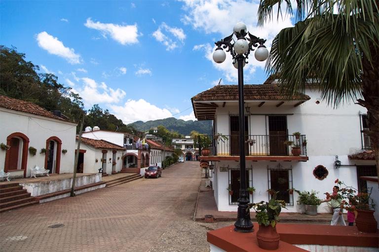 ¡Tapijulapa De Fiesta! Celebra 16 Años Como El Corazón Mágico De Tabasco 4 Una Pintoresca Calle Empedrada En El Pueblo Mágico De Tapijulapa, Tabasco, Flanqueada Por Casas De Arquitectura Virreinal Pintadas De Blanco Con Marcos Rojos, Tejados De Arcilla Marrón Y Balcones Decorados Con Flores. A La Derecha, Destaca Un Farol Público De Estilo Antiguo Con Cinco Esferas Blancas Junto A Una Palmera. Al Fondo, La Calle Adoquinada Sube Ligeramente Hacia Los Cerros Cubiertos De Densa Vegetación Bajo Un Cielo Azul Con Nubes Blancas.