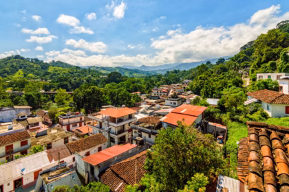 Vista Panorámica Desde Las Alturas Del Pueblo Mágico De Tapijulapa, Tabasco, Mostrando Un Pintoresco Conjunto De Casas Con Tejados De Teja De Arcilla Marrón Y Techos Rojizos, Enclavadas En Un Valle Rodeado Por Exuberantes Montañas Y Cerros Cubiertos De Densa Vegetación Selvática. Un Cielo Azul Brillante Salpicado De Nubes Blancas Cubre El Paisaje Serrano, Destacando La Armonía Entre La Arquitectura Tradicional Y La Naturaleza Del Sureste Mexicano.