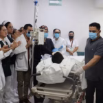 Fotografía En Plano Medio Horizontal Que Muestra Un Pasillo De Hospital Donde Personal Médico Y De Enfermería Forma Una Valla De Honor. Los Profesionales De La Salud, Portando Batas Blancas, Uniformes Quirúrgicos Y Cubrebocas, Aplauden Con Respeto Al Paso De Una Camilla De Hospital. En La Camilla, Equipada Con Un Soporte Para Soluciones Intravenosas, Es Trasladado Un Paciente Cubierto Con Sábanas Blancas. En Primer Plano A La Derecha, Un Enfermero Con Uniforme Gris Y Guantes Quirúrgicos Guía El Trayecto, Mientras Al Fondo Se Aprecia Un Letrero Verde De &Quot;Ruta De Evacuación&Quot;.