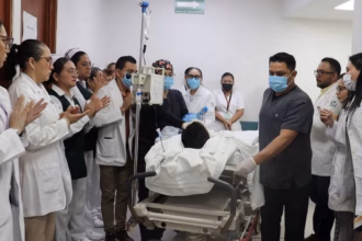 Fotografía En Plano Medio Horizontal Que Muestra Un Pasillo De Hospital Donde Personal Médico Y De Enfermería Forma Una Valla De Honor. Los Profesionales De La Salud, Portando Batas Blancas, Uniformes Quirúrgicos Y Cubrebocas, Aplauden Con Respeto Al Paso De Una Camilla De Hospital. En La Camilla, Equipada Con Un Soporte Para Soluciones Intravenosas, Es Trasladado Un Paciente Cubierto Con Sábanas Blancas. En Primer Plano A La Derecha, Un Enfermero Con Uniforme Gris Y Guantes Quirúrgicos Guía El Trayecto, Mientras Al Fondo Se Aprecia Un Letrero Verde De &Quot;Ruta De Evacuación&Quot;.
