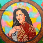 Fotografía En Primer Plano Y Formato Horizontal Que Muestra Un Mural Artístico Pintado Sobre Una Pared Exterior. La Obra Presenta El Retrato De La Reina De Belleza Fátima Bosch Enmarcado Dentro De Un Gran Círculo Con Textura De Madera. Ella Aparece Sonriendo, Con Cabello Largo Oscuro, Ojos Claros Y Portando Una Brillante Corona Dorada Sobre La Cabeza. Viste Un Elegante Vestido De Gala Color Rojo Oscuro Con Intrincados Bordados Dorados Y Destellos Blancos, Acompañado De Una Banda Plateada Cruzada Donde Se Lee Parcialmente La Palabra &Quot;Miss&Quot;. El Fondo Interior Del Círculo Combina Tonos Amarillos, Rosa Y Azul Celeste Pastel, Mientras Que Los Extremos Exteriores Del Mural Están Decorados Con Plantas Tropicales Verdes Y Una Gran Flor De Hibisco Roja.