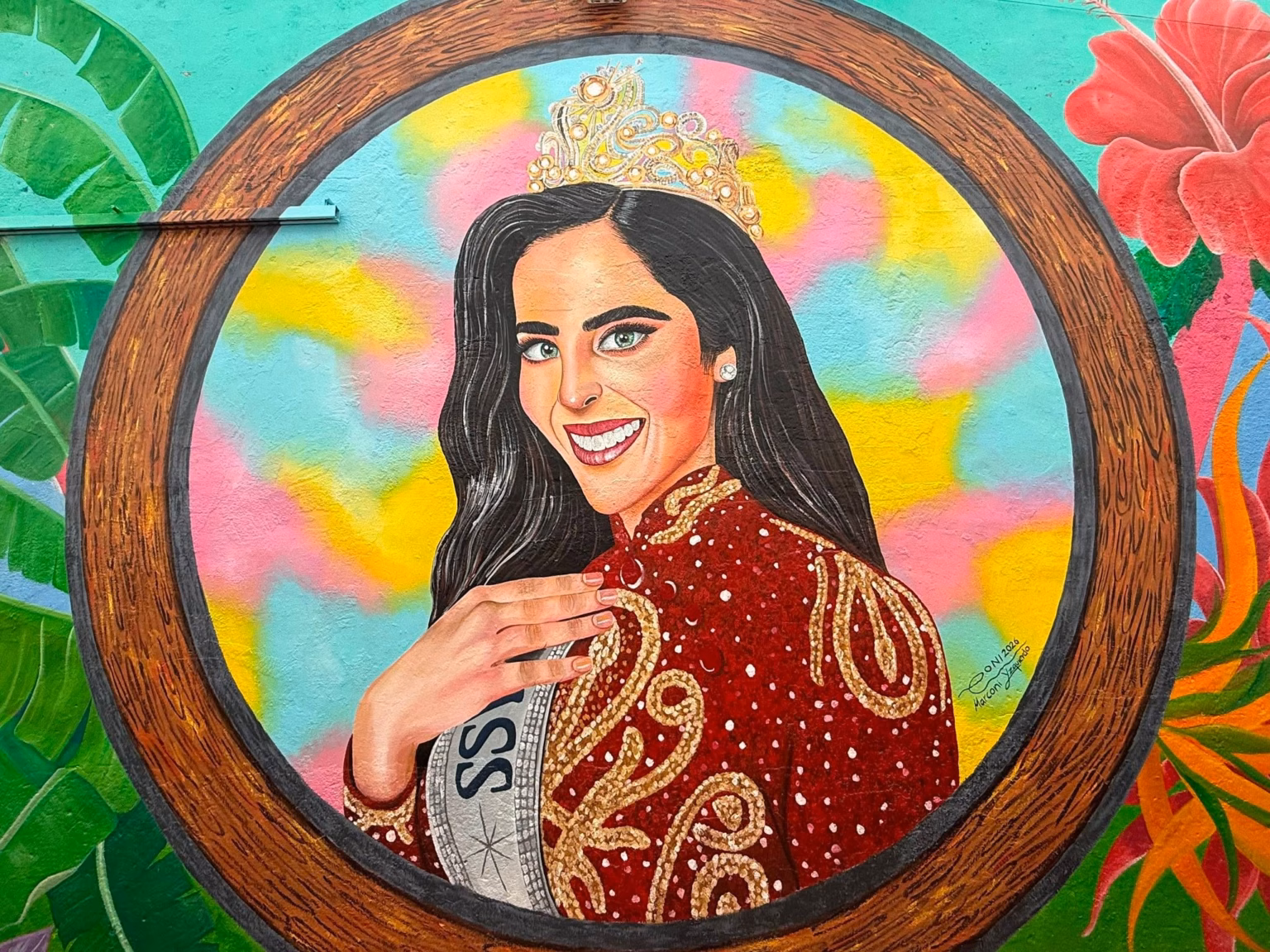 Fotografía En Primer Plano Y Formato Horizontal Que Muestra Un Mural Artístico Pintado Sobre Una Pared Exterior. La Obra Presenta El Retrato De La Reina De Belleza Fátima Bosch Enmarcado Dentro De Un Gran Círculo Con Textura De Madera. Ella Aparece Sonriendo, Con Cabello Largo Oscuro, Ojos Claros Y Portando Una Brillante Corona Dorada Sobre La Cabeza. Viste Un Elegante Vestido De Gala Color Rojo Oscuro Con Intrincados Bordados Dorados Y Destellos Blancos, Acompañado De Una Banda Plateada Cruzada Donde Se Lee Parcialmente La Palabra &Quot;Miss&Quot;. El Fondo Interior Del Círculo Combina Tonos Amarillos, Rosa Y Azul Celeste Pastel, Mientras Que Los Extremos Exteriores Del Mural Están Decorados Con Plantas Tropicales Verdes Y Una Gran Flor De Hibisco Roja.