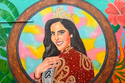 Fotografía En Primer Plano Y Formato Horizontal Que Muestra Un Mural Artístico Pintado Sobre Una Pared Exterior. La Obra Presenta El Retrato De La Reina De Belleza Fátima Bosch Enmarcado Dentro De Un Gran Círculo Con Textura De Madera. Ella Aparece Sonriendo, Con Cabello Largo Oscuro, Ojos Claros Y Portando Una Brillante Corona Dorada Sobre La Cabeza. Viste Un Elegante Vestido De Gala Color Rojo Oscuro Con Intrincados Bordados Dorados Y Destellos Blancos, Acompañado De Una Banda Plateada Cruzada Donde Se Lee Parcialmente La Palabra &Quot;Miss&Quot;. El Fondo Interior Del Círculo Combina Tonos Amarillos, Rosa Y Azul Celeste Pastel, Mientras Que Los Extremos Exteriores Del Mural Están Decorados Con Plantas Tropicales Verdes Y Una Gran Flor De Hibisco Roja.