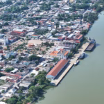 Vista Aérea Panorámica De La Ciudad Costera Y Puerto De Frontera, Tabasco. A La Derecha, Se Observa La Amplia Desembocadura De Un Río De Tonalidades Verdosas Con Embarcaciones Comerciales Atracadas Junto Al Malecón. A La Izquierda, Se Extiende El Trazado Urbano Con Casas De Techos Diversos, Abundante Vegetación Tropical Interconectada Entre Las Cuadras, La Plaza Central Con Su Quiosco Y La Estructura Blanca De La Iglesia Parroquial Destacando Entre Los Edificios Locales.