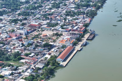 Vista Aérea Panorámica De La Ciudad Costera Y Puerto De Frontera, Tabasco. A La Derecha, Se Observa La Amplia Desembocadura De Un Río De Tonalidades Verdosas Con Embarcaciones Comerciales Atracadas Junto Al Malecón. A La Izquierda, Se Extiende El Trazado Urbano Con Casas De Techos Diversos, Abundante Vegetación Tropical Interconectada Entre Las Cuadras, La Plaza Central Con Su Quiosco Y La Estructura Blanca De La Iglesia Parroquial Destacando Entre Los Edificios Locales.