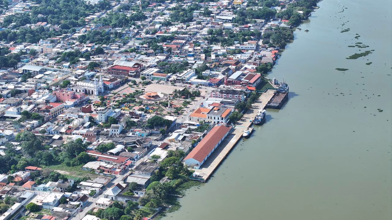 Vista Aérea Panorámica De La Ciudad Costera Y Puerto De Frontera, Tabasco. A La Derecha, Se Observa La Amplia Desembocadura De Un Río De Tonalidades Verdosas Con Embarcaciones Comerciales Atracadas Junto Al Malecón. A La Izquierda, Se Extiende El Trazado Urbano Con Casas De Techos Diversos, Abundante Vegetación Tropical Interconectada Entre Las Cuadras, La Plaza Central Con Su Quiosco Y La Estructura Blanca De La Iglesia Parroquial Destacando Entre Los Edificios Locales.
