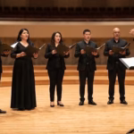 Un Plano Horizontal Medio-Largo De Un Coro Mixto De 10 Personas Y Un Director Actuando En Un Gran Auditorio Vacío. Diez Cantantes (Cuatro Mujeres Y Seis Hombres), Vestidos De Negro Formal De Concierto Y Sosteniendo Carpetas De Partituras Abiertas, Están De Pie En Formación De Semicírculo En Un Escenario De Madera.