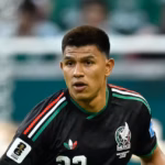 El Futbolista Mexicano Jesús Gallardo En Plena Acción Sobre El Césped Durante Un Partido De Fútbol. Viste El Uniforme Negro De La Selección Mexicana Con El Número 23 En El Pecho Y En El Short, Detalles Tricolores En Los Hombros, Calcetas Negras Altas Y Tacos De Color Rosa Brillante. Su Mirada Está Fija Al Frente, Manteniendo Una Postura Alerta Cerca Del Balón, El Cual Se Observa En El Pasto A La Esquina Inferior Derecha. Al Fondo Desenfocado Se Aprecian Las Tribunas Y Una Portería.