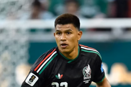 El Futbolista Mexicano Jesús Gallardo En Plena Acción Sobre El Césped Durante Un Partido De Fútbol. Viste El Uniforme Negro De La Selección Mexicana Con El Número 23 En El Pecho Y En El Short, Detalles Tricolores En Los Hombros, Calcetas Negras Altas Y Tacos De Color Rosa Brillante. Su Mirada Está Fija Al Frente, Manteniendo Una Postura Alerta Cerca Del Balón, El Cual Se Observa En El Pasto A La Esquina Inferior Derecha. Al Fondo Desenfocado Se Aprecian Las Tribunas Y Una Portería.