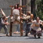 Uatro Actores Actúan En Una Obra De Teatro Callejero En Un Parque. Un Actor Lleva Una Estructura De Madera Con Un Panel De Madera, Una Actriz Lleva Un Disfraz De Mujer Gorda, Un Actor Lleva Un Disfraz De Granjero Y Una Actriz Lleva Un Vestido Con Capas Y Tiene Los Brazos Extendidos. Hay Público Sentado Detrás De Ellos.