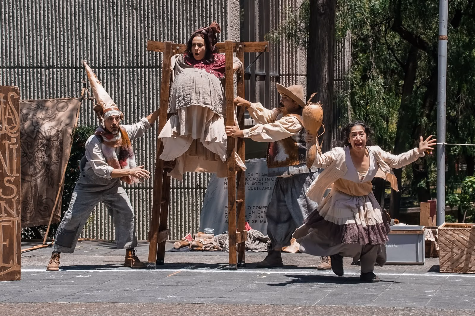 ¿Dónde Ir Este Viernes? El Festival Ceiba Trae A Plaza De Armas El Imperdible Espectáculo Callejero ¡Burros! 72 Uatro Actores Actúan En Una Obra De Teatro Callejero En Un Parque. Un Actor Lleva Una Estructura De Madera Con Un Panel De Madera, Una Actriz Lleva Un Disfraz De Mujer Gorda, Un Actor Lleva Un Disfraz De Granjero Y Una Actriz Lleva Un Vestido Con Capas Y Tiene Los Brazos Extendidos. Hay Público Sentado Detrás De Ellos.