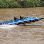 Una Lancha De Carreras Azul Con El Número 372 Y Un Motor Fuera De Borda Mercury Navega A Gran Velocidad Sobre El Agua Color Café Del Río Grijalva. Dos Tripulantes Con Cascos De Protección Van A Bordo Mientras La Embarcación Deja Una Estela De Agua Tras De Sí, Con Vegetación Verde Visible Al Fondo En La Orilla Del Río.