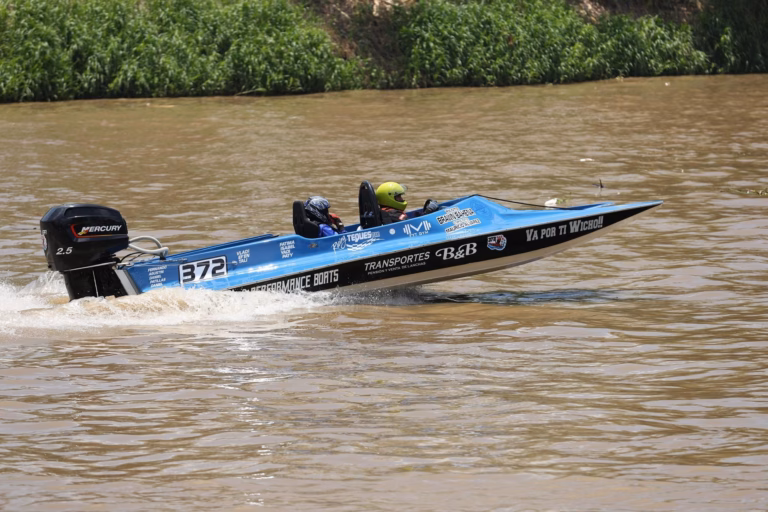 Una Lancha De Carreras Azul Con El Número 372 Y Un Motor Fuera De Borda Mercury Navega A Gran Velocidad Sobre El Agua Color Café Del Río Grijalva. Dos Tripulantes Con Cascos De Protección Van A Bordo Mientras La Embarcación Deja Una Estela De Agua Tras De Sí, Con Vegetación Verde Visible Al Fondo En La Orilla Del Río.
