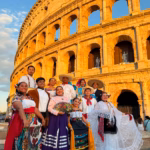 Un Grupo De Nueve Bailarines Mexicanos Del Ballet &Quot;Winik Asik Ak'Ot&Quot;, Vestidos Con Coloridos Trajes Folclóricos Tradicionales De Tabasco (Incluyendo Faldas Bordadas, Vestidos De Encaje Blanco, Camisas Blancas Y Sombreros De Ala Ancha), Posan Sonrientes Frente Al Coliseo De Roma Al Atardecer.