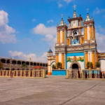 Fachada Colorida E Icónica De La Iglesia De La Virgen De La Asunción En Cupilco, Comalcalco, Pintada En Tonos Vivos De Amarillo, Azul, Verde Y Blanco, Con Una Amplia Plaza Frontal Bajo Un Cielo Despejado.