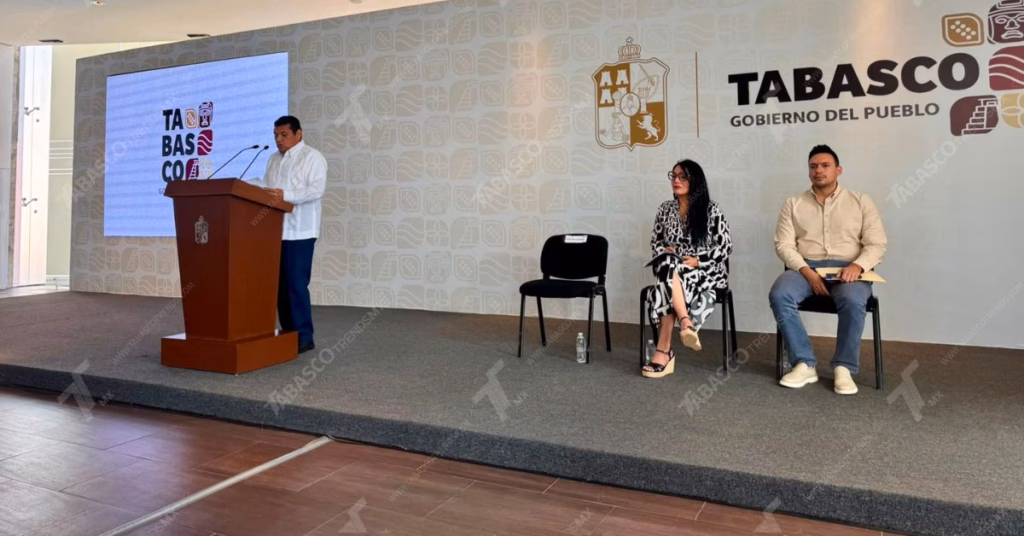 El Gobernador De Tabasco, Javier May, De Pie Detrás De Un Podio, Hablando Por Micrófonos. Viste Una Guayabera Blanca Y Pantalones Oscuros. Detrás De Él Hay Una Gran Pantalla Con Gráficos Y Texto, Que Incluye El Logo &Quot;Tabasco&Quot; Y &Quot;Gobierno Del Pueblo&Quot;. A Su Derecha, Una Mujer Con Un Vestido Estampado Y Un Hombre Con Una Camisa Beige Están Sentados En Sillas. La Sala Es Moderna Con Grandes Ventanales. La Marca De Agua &Quot;Tabasco&Quot; Está Visible En La Imagen.