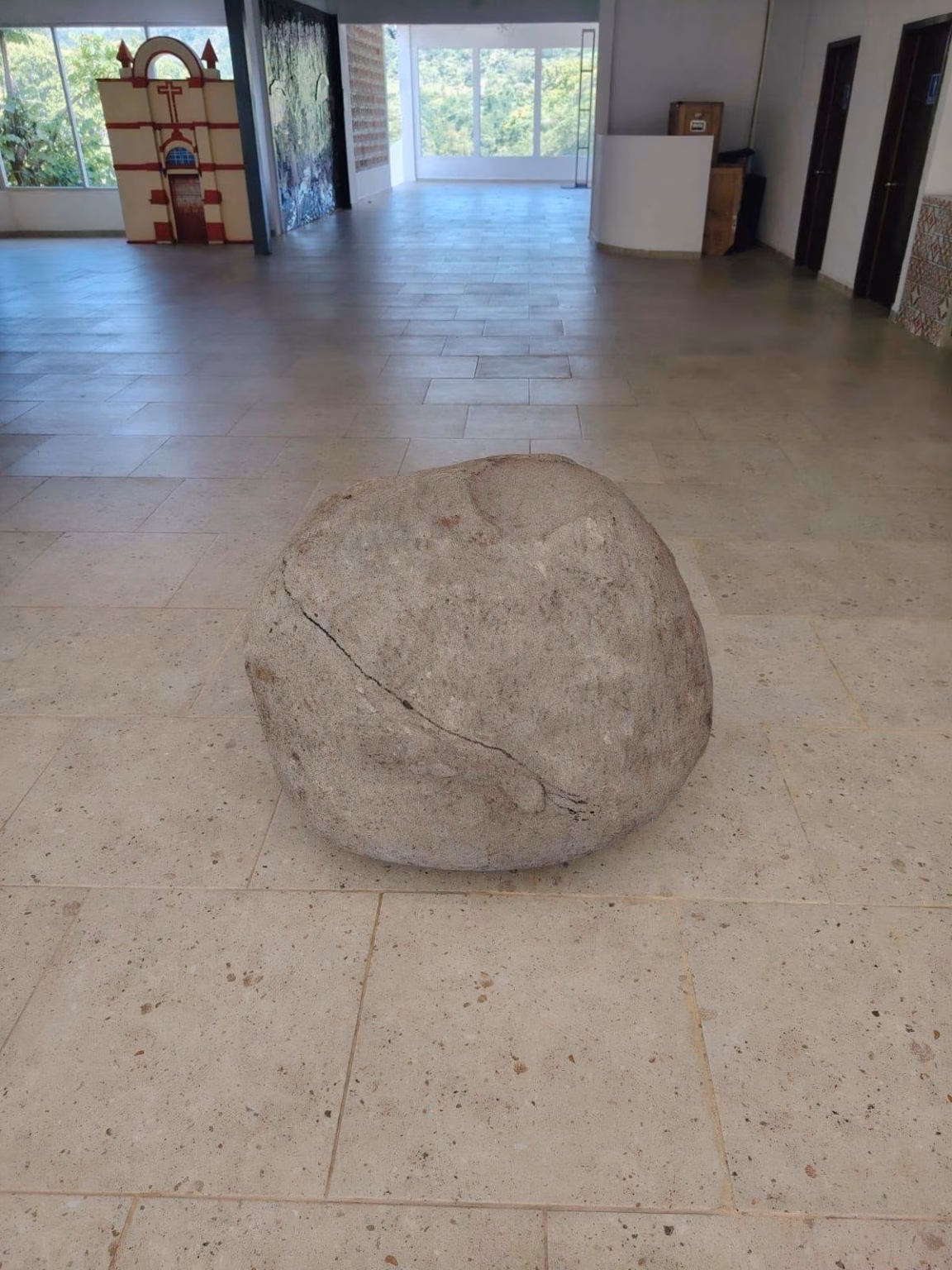 Una Gran Roca Esférica De Piedra Caliza, Con Una Fisura Diagonal, Exhibida En El Centro De Una Sala De Museo Sobre Un Piso De Losas Claras. Al Fondo, A La Izquierda, Se Observa Una Maqueta De La Fachada De Una Iglesia, Y A La Derecha, Una Ventana Que Da Al Exterior Y Puertas De Madera.
