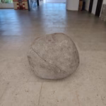 Una Gran Roca Esférica De Piedra Caliza, Con Una Fisura Diagonal, Exhibida En El Centro De Una Sala De Museo Sobre Un Piso De Losas Claras. Al Fondo, A La Izquierda, Se Observa Una Maqueta De La Fachada De Una Iglesia, Y A La Derecha, Una Ventana Que Da Al Exterior Y Puertas De Madera.