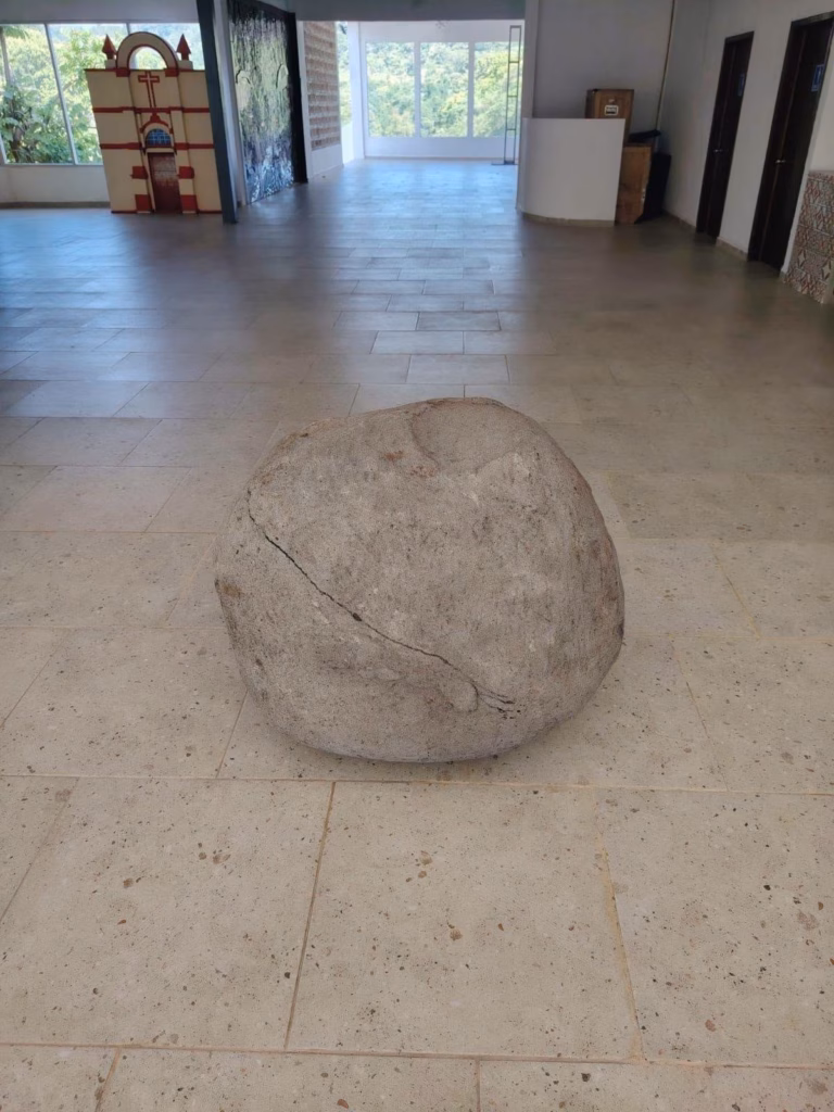 Una Gran Roca Esférica De Piedra Caliza, Con Una Fisura Diagonal, Exhibida En El Centro De Una Sala De Museo Sobre Un Piso De Losas Claras. Al Fondo, A La Izquierda, Se Observa Una Maqueta De La Fachada De Una Iglesia, Y A La Derecha, Una Ventana Que Da Al Exterior Y Puertas De Madera.