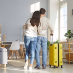 Una Pareja Abraza De Espaldas Contemplando Su Sala Limpia Y Ordenada Junto A Una Maleta Amarilla, Lista Para Preparar Tu Casa En Tabasco Antes De Salir De Vacaciones.