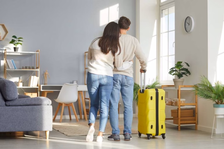 Una Pareja Abraza De Espaldas Contemplando Su Sala Limpia Y Ordenada Junto A Una Maleta Amarilla, Lista Para Preparar Tu Casa En Tabasco Antes De Salir De Vacaciones.