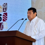 Otografía En Plano Medio Del Gobernador Javier May Rodríguez De Pie En Un Podio, Hablando Por Dos Micrófonos. Lleva Una Guayabera Blanca Bordada. Detrás De Él, Una Gran Pantalla Azul Muestra Logotipos De Estilo Gráfico Prehispánico En Blanco, Marrón Y Naranja, Y Parte De La Palabra &Quot;Tabasco&Quot; Es Visible. Javier May Mira Hacia La Derecha Mientras Habla.
