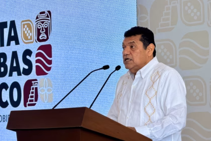 Otografía En Plano Medio Del Gobernador Javier May Rodríguez De Pie En Un Podio, Hablando Por Dos Micrófonos. Lleva Una Guayabera Blanca Bordada. Detrás De Él, Una Gran Pantalla Azul Muestra Logotipos De Estilo Gráfico Prehispánico En Blanco, Marrón Y Naranja, Y Parte De La Palabra &Quot;Tabasco&Quot; Es Visible. Javier May Mira Hacia La Derecha Mientras Habla.